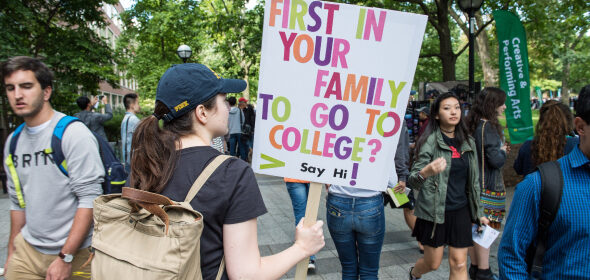 A student walks through campus holding a sign asking passers-by if they are the first person in their family to go to college