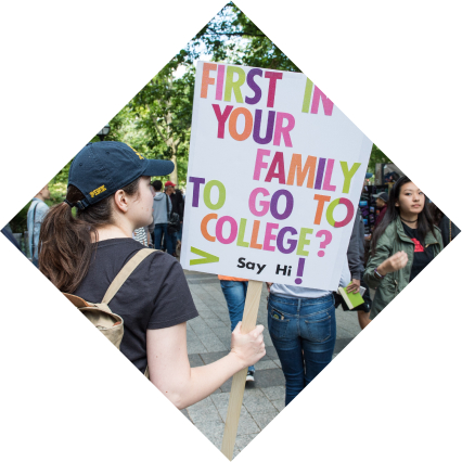 A student walks through campus holding a poster which reads "First in your family to go to college?"