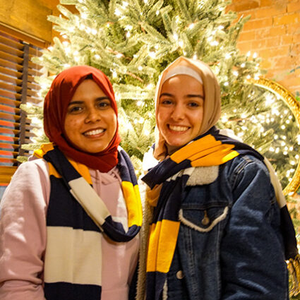 Two smiling women in hijabs with matching maize, blue, and white scarves around their necks