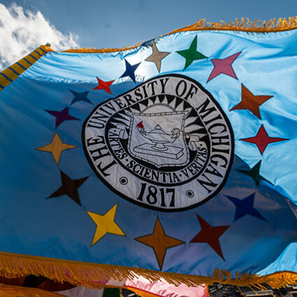 A U-M flag with the seal on it and multi-colored stars flying against a bright blue sky