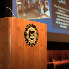 A wooden podium with the U-M seal on it and a presentation being projected on a screen behind it